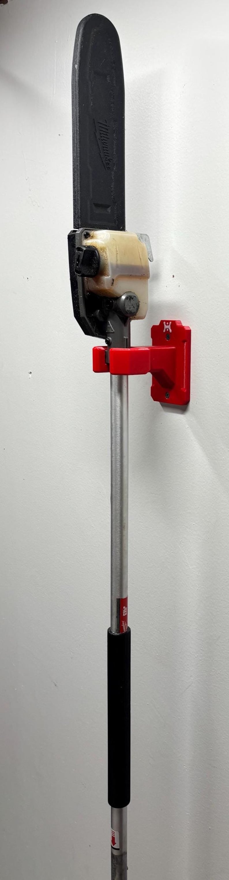 May include: A pole saw mounted on a wall. The saw has a black blade, a tan engine housing, and a silver pole with a black handle. A red bracket holds the saw to the wall.