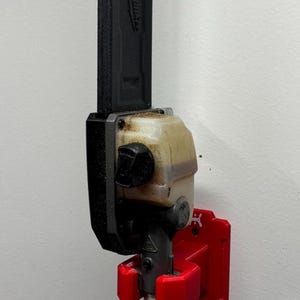 May include: A black and silver pole saw with a red mounting bracket attached to a white wall. The pole saw is labeled "Milwaukee". The mounting bracket is red.