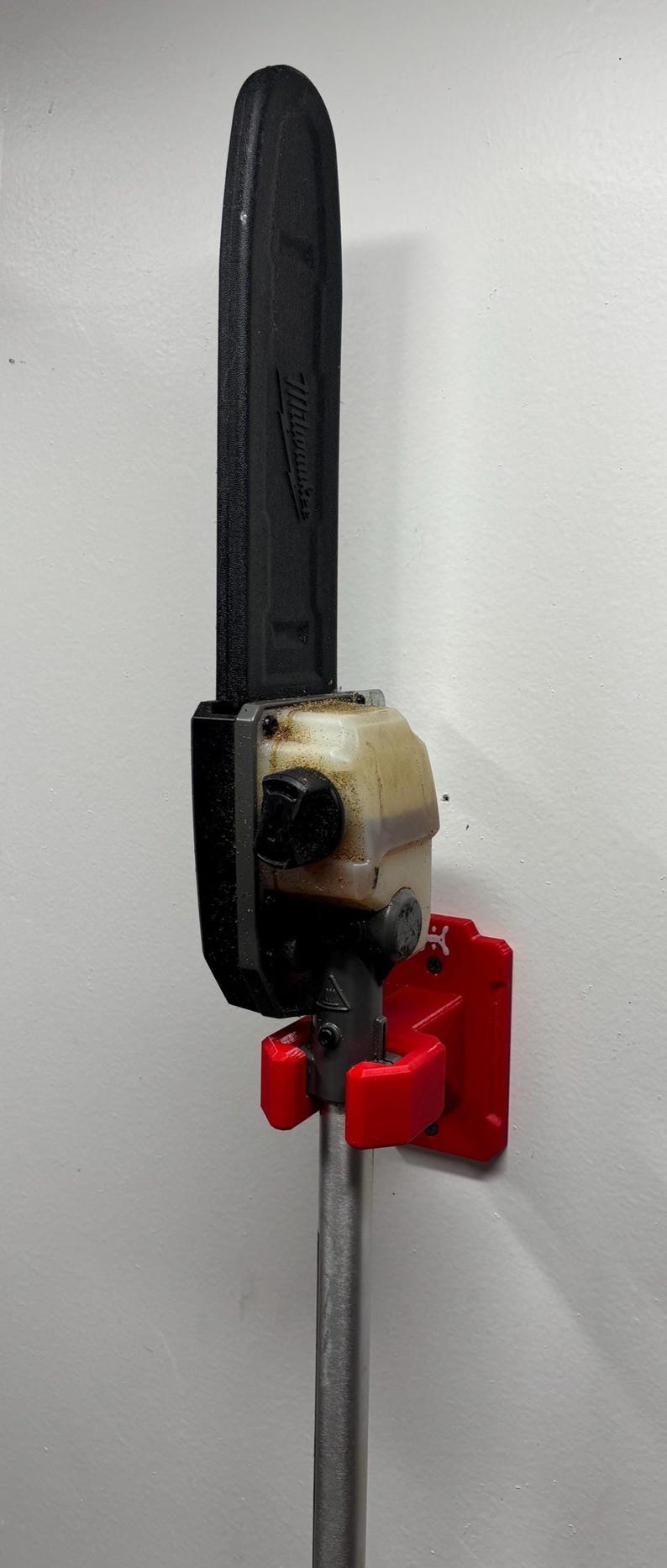 May include: A black and silver pole saw with a red mounting bracket attached to a white wall. The pole saw is labeled "Milwaukee". The mounting bracket is red.