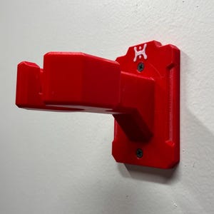 May include: A bright red wall-mounted bracket, likely 3D-printed, with a rectangular base and a projecting arm. The bracket is secured to a white wall with two screws. A small white logo is visible on the top of the bracket.