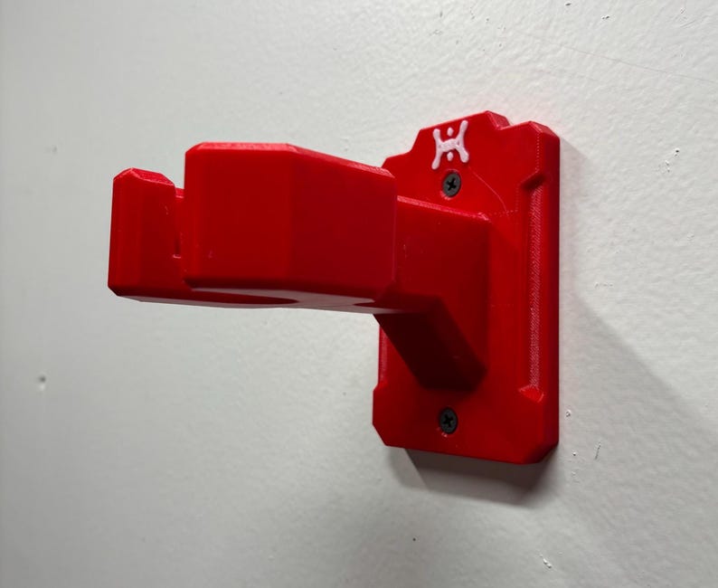 May include: A bright red wall-mounted bracket, likely 3D-printed, with a rectangular base and a projecting arm. The bracket is secured to a white wall with two screws. A small white logo is visible on the top of the bracket.