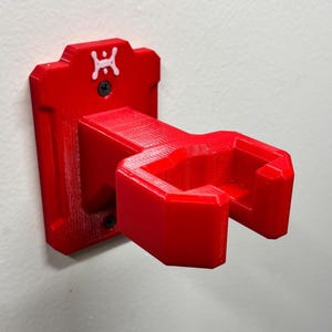 May include: A bright red, 3D-printed wall-mounted holder. The holder features a rectangular backplate with a small white logo and two screw holes. The angled cradle is designed to hold an item. The holder is mounted on a white wall.