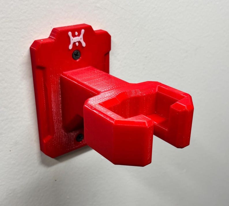 May include: A bright red, 3D-printed wall-mounted holder. The holder features a rectangular backplate with a small white logo and two screw holes. The angled cradle is designed to hold an item. The holder is mounted on a white wall.