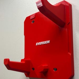 Puede incluir: Un soporte de pared rojo brillante con dos brazos salientes para apoyo. La base rectangular tiene la palabra "EVERSION" grabada en letras blancas en el centro. Se observan tres tornillos negros que fijan el soporte a la pared.