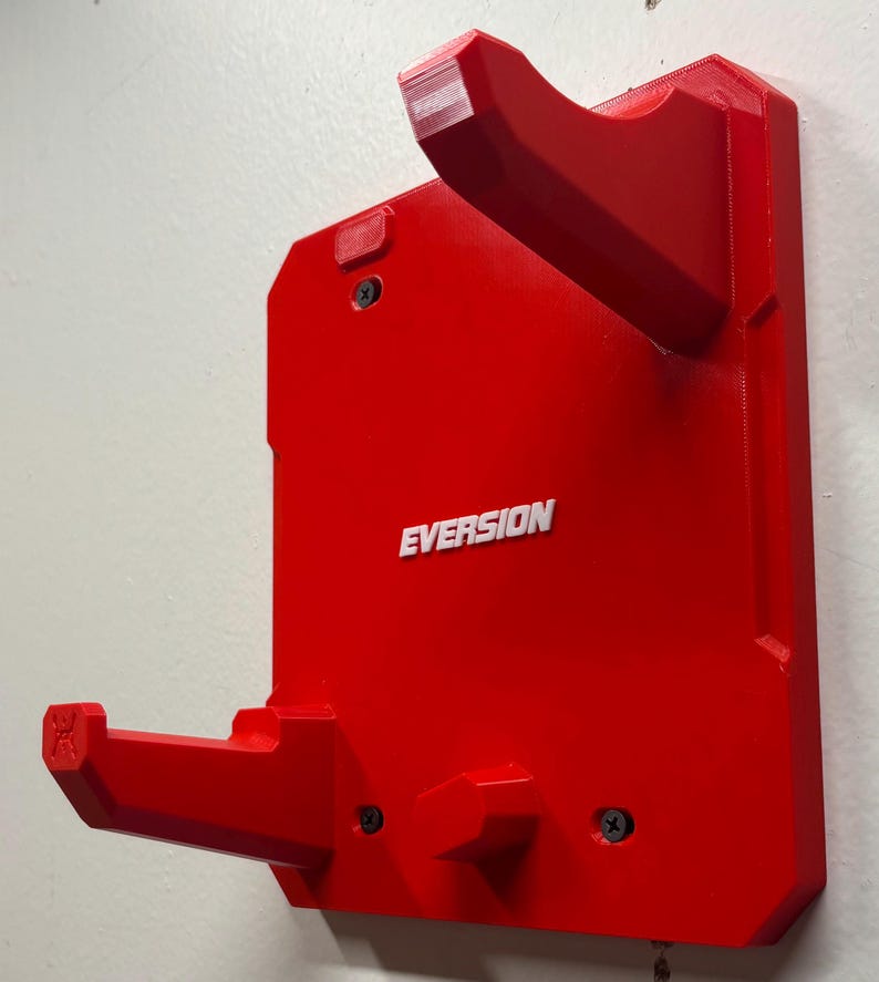 Puede incluir: Un soporte de pared rojo brillante con dos brazos salientes para apoyo. La base rectangular tiene la palabra "EVERSION" grabada en letras blancas en el centro. Se observan tres tornillos negros que fijan el soporte a la pared.