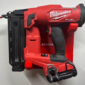 Puede incluir: Una clavadora inal&aacute;mbrica Milwaukee Fuel roja y negra montada en un soporte de pared de pl&aacute;stico rojo. La herramienta cuenta con un cargador negro, una empu&ntilde;adura texturizada y una bater&iacute;a M18 Redlithium CP2.0 con un gancho de cintur&oacute;n met&aacute;lico.
