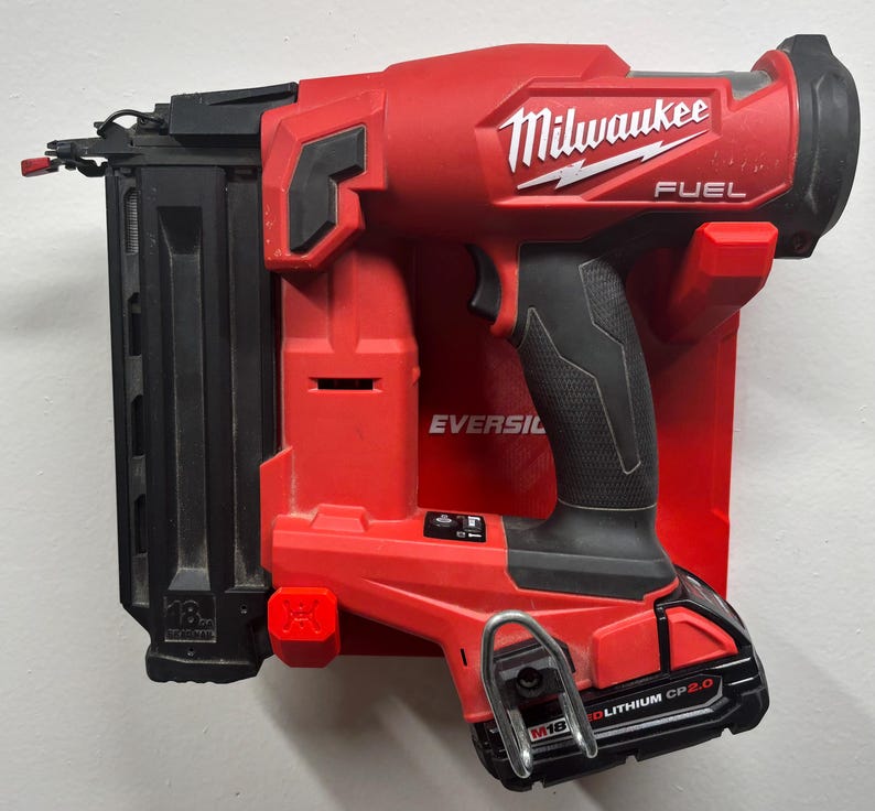 Puede incluir: Una clavadora inal&aacute;mbrica Milwaukee Fuel roja y negra montada en un soporte de pared de pl&aacute;stico rojo. La herramienta cuenta con un cargador negro, una empu&ntilde;adura texturizada y una bater&iacute;a M18 Redlithium CP2.0 con un gancho de cintur&oacute;n met&aacute;lico.