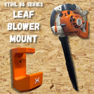 May include: An orange leaf blower mount and a leaf blower are displayed against a wood-grain background. The mount is a bright orange, with the text "STIHL 86 SERIES LEAF BLOWER MOUNT" in white. The leaf blower is orange, black, and white.