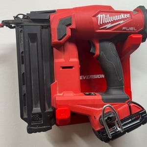 Puede incluir: Una clavadora inal&aacute;mbrica Milwaukee Fuel de calibre 18 en color rojo y negro montada en un soporte de pared de pl&aacute;stico rojo. La herramienta cuenta con un cargador negro, empu&ntilde;adura de goma negra y bater&iacute;a M18 Lithium CP2.0 con gancho met&aacute;lico.