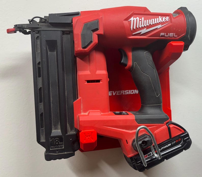 Puede incluir: Una clavadora inal&aacute;mbrica Milwaukee Fuel de calibre 18 en color rojo y negro montada en un soporte de pared de pl&aacute;stico rojo. La herramienta cuenta con un cargador negro, empu&ntilde;adura de goma negra y bater&iacute;a M18 Lithium CP2.0 con gancho met&aacute;lico.