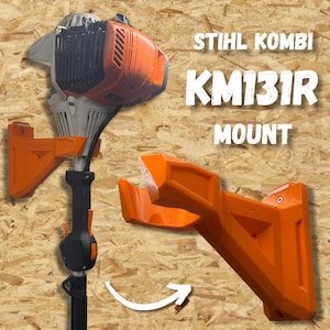 May include: An orange wall-mounted bracket designed for a Stihl Kombi KM131R power tool. The bracket holds the tool shaft securely. The text 'STIHL KOMBI KM131R MOUNT' is visible. The mount features a rigid, angular design with screw holes.
