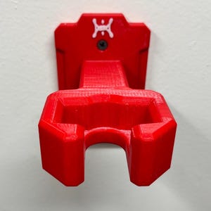 May include: A bright red, 3D-printed wall mount. The mount features a bracket with a curved opening and a rectangular top with a white logo and a screw. The mount is designed to hold an item, and is mounted on a white wall.