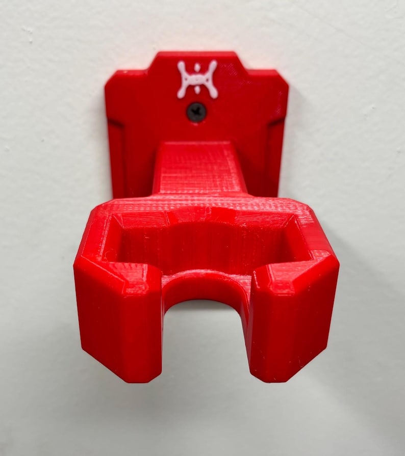 May include: A bright red, 3D-printed wall mount. The mount features a bracket with a curved opening and a rectangular top with a white logo and a screw. The mount is designed to hold an item, and is mounted on a white wall.