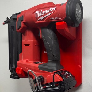 Puede incluir: Una clavadora inal&aacute;mbrica roja Milwaukee Fuel montada en un soporte de pared de pl&aacute;stico rojo. La herramienta cuenta con un mango negro, un cargador de clavos negro y una bater&iacute;a negra RedLithium CP 2.0 en la base con un gancho de metal plateado.