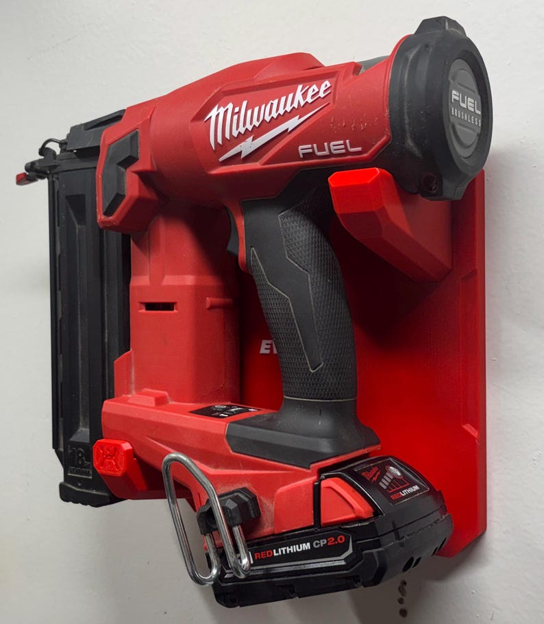 Puede incluir: Una clavadora inal&aacute;mbrica roja Milwaukee Fuel montada en un soporte de pared de pl&aacute;stico rojo. La herramienta cuenta con un mango negro, un cargador de clavos negro y una bater&iacute;a negra RedLithium CP 2.0 en la base con un gancho de metal plateado.