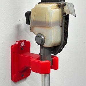 May include: A red wall mount for a pole saw, holding a black and silver pole saw. The mount is red and has a logo on it. The pole saw has a black blade guard and a clear fuel tank.