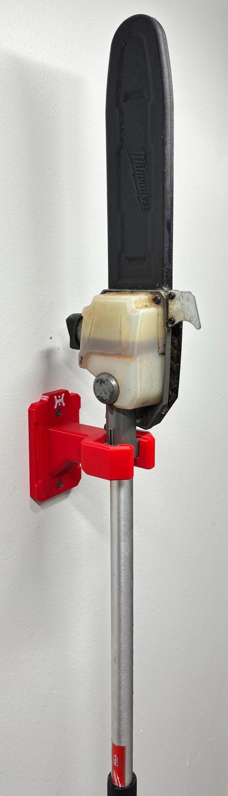 May include: A red wall mount for a pole saw, holding a black and silver pole saw. The mount is red and has a logo on it. The pole saw has a black blade guard and a clear fuel tank.
