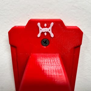 May include: Close-up of a bright red, geometric wall mount with a white emblem and a black screw. The mount has a textured surface and a curved bottom, designed for holding an item.