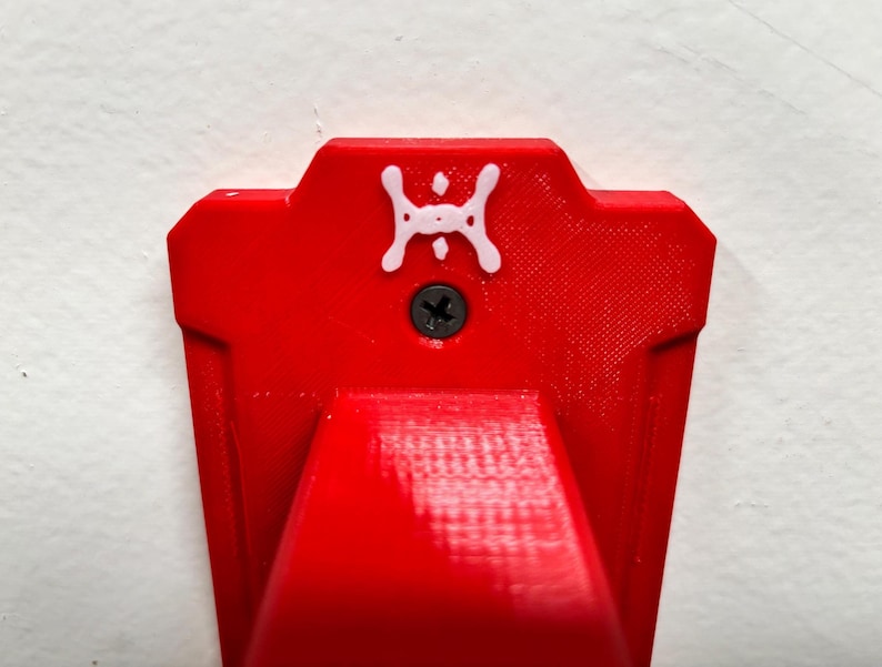 May include: Close-up of a bright red, geometric wall mount with a white emblem and a black screw. The mount has a textured surface and a curved bottom, designed for holding an item.