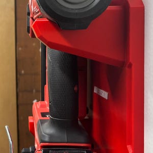 Puede incluir: Un soporte de pared rojo para herramientas que sostiene una herramienta el&eacute;ctrica Milwaukee Fuel Brushless. La herramienta cuenta con un mango negro texturizado, una bater&iacute;a etiquetada como Milwaukee Redlithium con indicador de carga y un gancho de metal plateado.