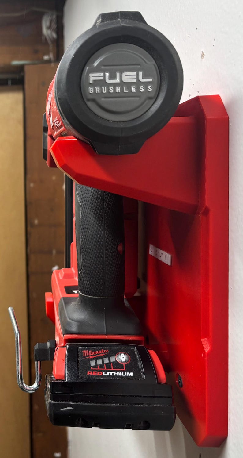 Puede incluir: Un soporte de pared rojo para herramientas que sostiene una herramienta el&eacute;ctrica Milwaukee Fuel Brushless. La herramienta cuenta con un mango negro texturizado, una bater&iacute;a etiquetada como Milwaukee Redlithium con indicador de carga y un gancho de metal plateado.