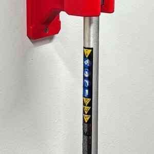 May include: A black pole saw with a red mounting bracket. The saw blade is black, and the pole is silver with a black grip. The saw is mounted on a white wall.