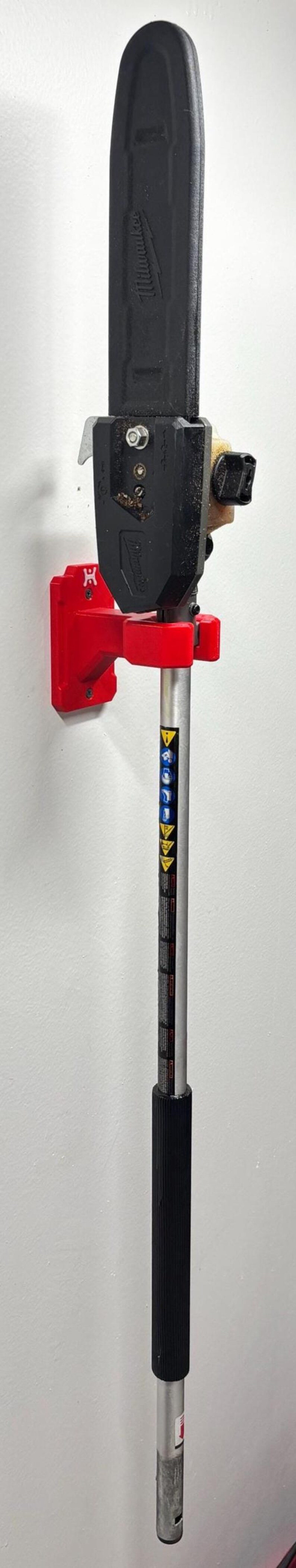 May include: A black pole saw with a red mounting bracket. The saw blade is black, and the pole is silver with a black grip. The saw is mounted on a white wall.