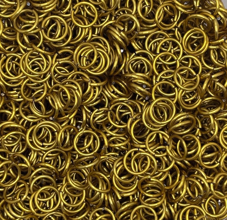 May include: A large collection of small, circular gold-colored metal jump rings. Each individual ring is uniform in shape and size, featuring a smooth, polished finish. The rings are densely packed together, creating a textured, metallic surface.