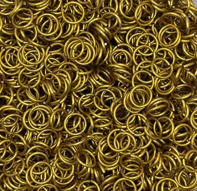 Puede incluir: Una gran colecci&oacute;n de peque&ntilde;os anillos de salto met&aacute;licos circulares de color dorado. Cada anillo es uniforme en forma y tama&ntilde;o, con un acabado liso y pulido. Los anillos est&aacute;n densamente agrupados, creando una superficie met&aacute;lica texturizada.