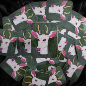 May include: A collection of square stickers featuring a white deer head with pink ears and a pink nose, set against a green background. The deer has yellow antlers and a tear. The stickers are scattered on a black surface.