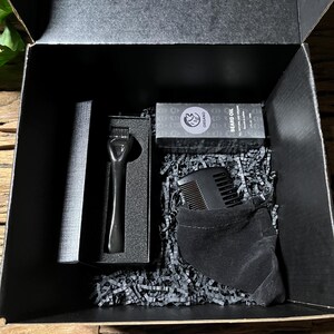 May include: A black gift box with black shredded paper inside. The box contains a black beard comb, a black beard oil bottle with the text "BEARD OIL" and a black pouch.