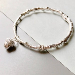 May include: A silver bangle bracelet with a textured twig design and an acorn charm.