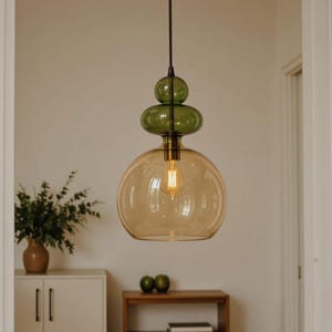 Op de afbeelding: Een moderne hanglamp met een gelaagd ontwerp. De lamp heeft een grote, amberkleurige glazen bol aan de onderkant, bekroond door een groen glazen gedeelte. Een enkele gloeilamp is zichtbaar in de bol. De lamp hangt aan een zwart snoer.
