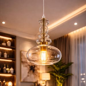 Puede incluir: Una lámpara colgante de vidrio transparente con un vástago de metal dorado y una gran pantalla de vidrio redondeada. La bombilla es visible dentro de la pantalla, emitiendo un brillo cálido y amarillo. La lámpara cuelga de un cable blanco.
