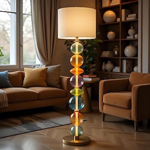 Puede incluir: Una lámpara de pie moderna con una pantalla blanca y una base de orbes de vidrio de colores. La base presenta esferas apiladas en tonos de azul, verde, naranja y morado. La lámpara está en un salón con un sofá y un sillón marrones.