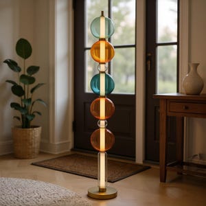 Puede incluir: Una lámpara de pie moderna con base de latón y una serie de globos de vidrio de colores en tonos azul, naranja y transparente. La lámpara está situada en una habitación bien iluminada con una paleta de colores neutros.