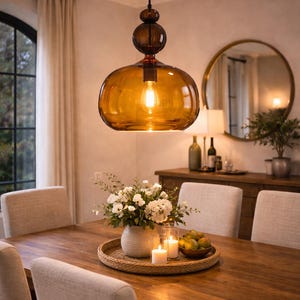 Puede incluir: Una lámpara colgante con una pantalla de cristal ámbar cuelga sobre una mesa de comedor de madera. La luz está encendida, creando un ambiente cálido. La mesa está decorada con flores, velas y fruta. La habitación tiene una paleta de colores neutros.