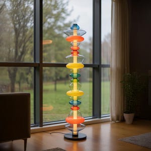 May include: A tall, colorful floor lamp with a black base. The lamp features stacked, translucent, disc-shaped elements in shades of blue, orange, yellow, and green. The lamp is positioned near a large window, with a view of trees and a cloudy sky.