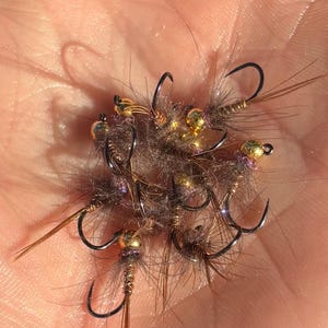 May include: A close-up of several fishing flies. The flies have black hooks, gold-colored beads, and brown and gray feathery bodies. The flies are held in a person's hand.