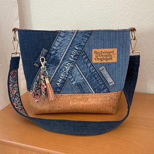 Peut inclure: Sac bandoulière en jean avec une base en liège et une sangle à motifs floraux. Le sac porte les inscriptions "AMERICAN EAGLE" et "Reclaimed Denim Originals". Une pompon et un charm pendent de la fermeture éclair.