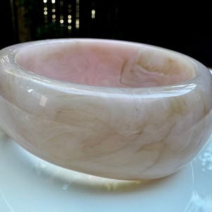 May include: A wide, round bangle bracelet in shades of pink and cream. The bracelet has a marbled appearance, with swirling patterns throughout. The smooth, polished surface reflects light, creating a subtle sheen. This accessory is a statement piece.