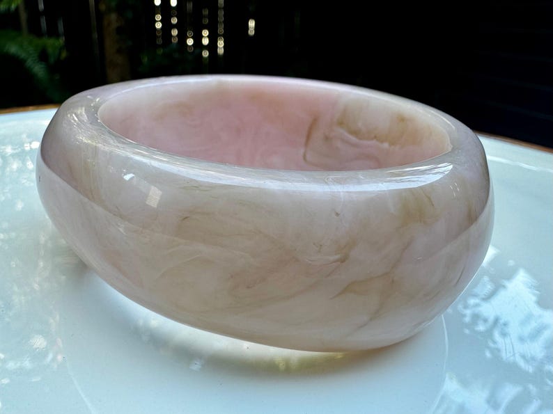 May include: A wide, round bangle bracelet in shades of pink and cream. The bracelet has a marbled appearance, with swirling patterns throughout. The smooth, polished surface reflects light, creating a subtle sheen. This accessory is a statement piece.