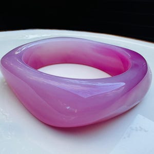 May include: A translucent, square-shaped bangle in shades of lavender and magenta. The smooth, polished surface reflects light, highlighting the variations in color. This piece of jewelry is a statement accessory.
