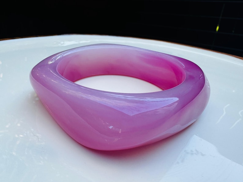 May include: A translucent, square-shaped bangle in shades of lavender and magenta. The smooth, polished surface reflects light, highlighting the variations in color. This piece of jewelry is a statement accessory.