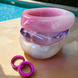 May include: A stack of three large, translucent, pastel-colored bangles in shades of pink, purple, and white. Two smaller, bright pink rings are in front of the stack. The jewelry is displayed on a light brown surface with a pool and greenery in the background.