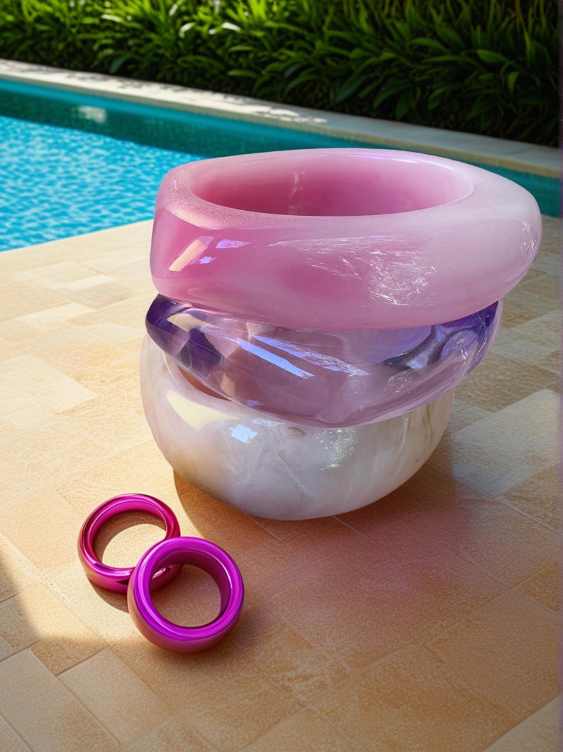 May include: A stack of three large, translucent, pastel-colored bangles in shades of pink, purple, and white. Two smaller, bright pink rings are in front of the stack. The jewelry is displayed on a light brown surface with a pool and greenery in the background.