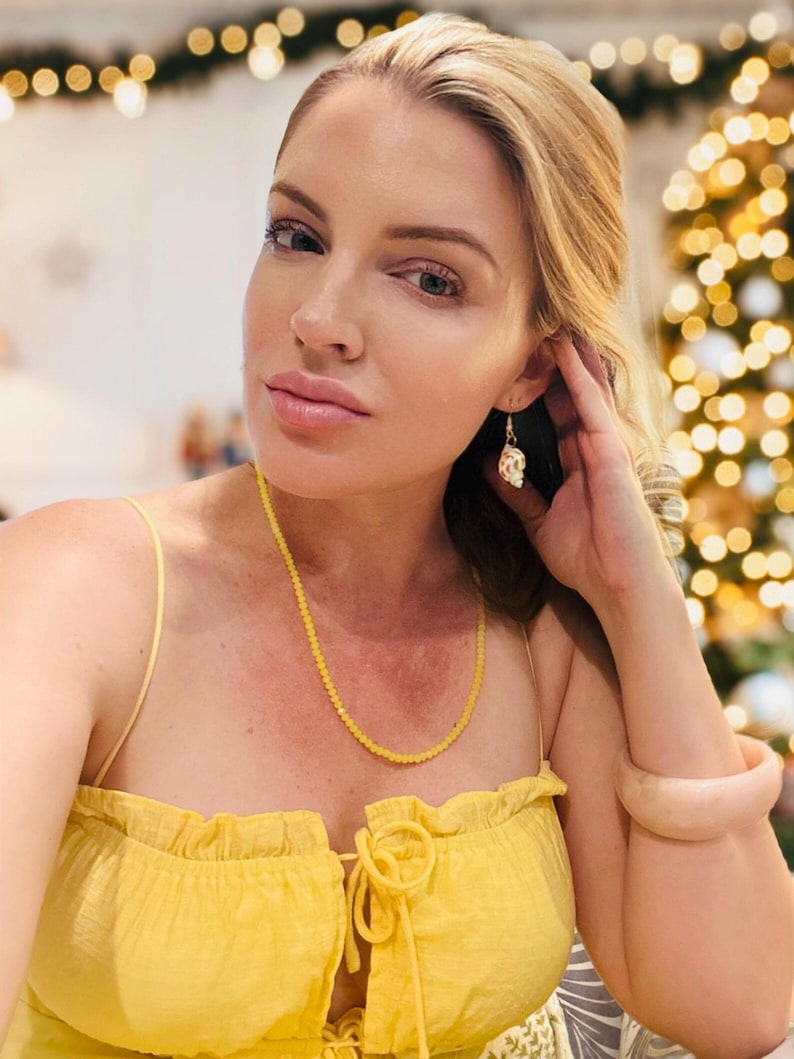 May include: A woman wearing a yellow top, a beaded yellow necklace, and a light pink bracelet. The earrings are a gold color with a white design. The background is blurred with Christmas lights.