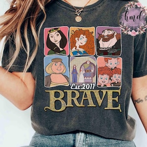 May include: Dark gray t-shirt featuring a graphic of characters from the animated film "Brave." The design includes six framed images of characters, the text "Est. 2011," and the word "BRAVE" in gold lettering.