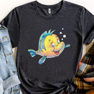 K&ouml;nnte beinhalten: Dunkelgraues T-Shirt mit einer Cartoon-Illustration eines gelb-blauen Fisches, der l&auml;chelt und Blasen hat. Der Fisch hat blaue Augen und schwimmt nach rechts. Das T-Shirt ist aus weichem Material.