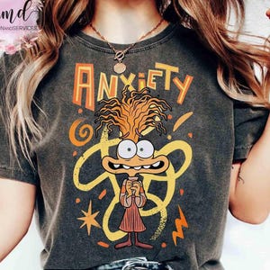 Peut inclure: T-shirt gris foncé avec le mot "ANXIETY" en orange au-dessus d'un personnage de dessin animé aux cheveux orange et à l'expression inquiète. Le motif comprend des tourbillons, des étoiles et des éclairs jaunes.